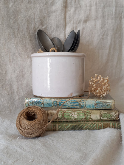 Antique jam pot on a textured surface containing spoons, dried flowers, and books on a textured surface