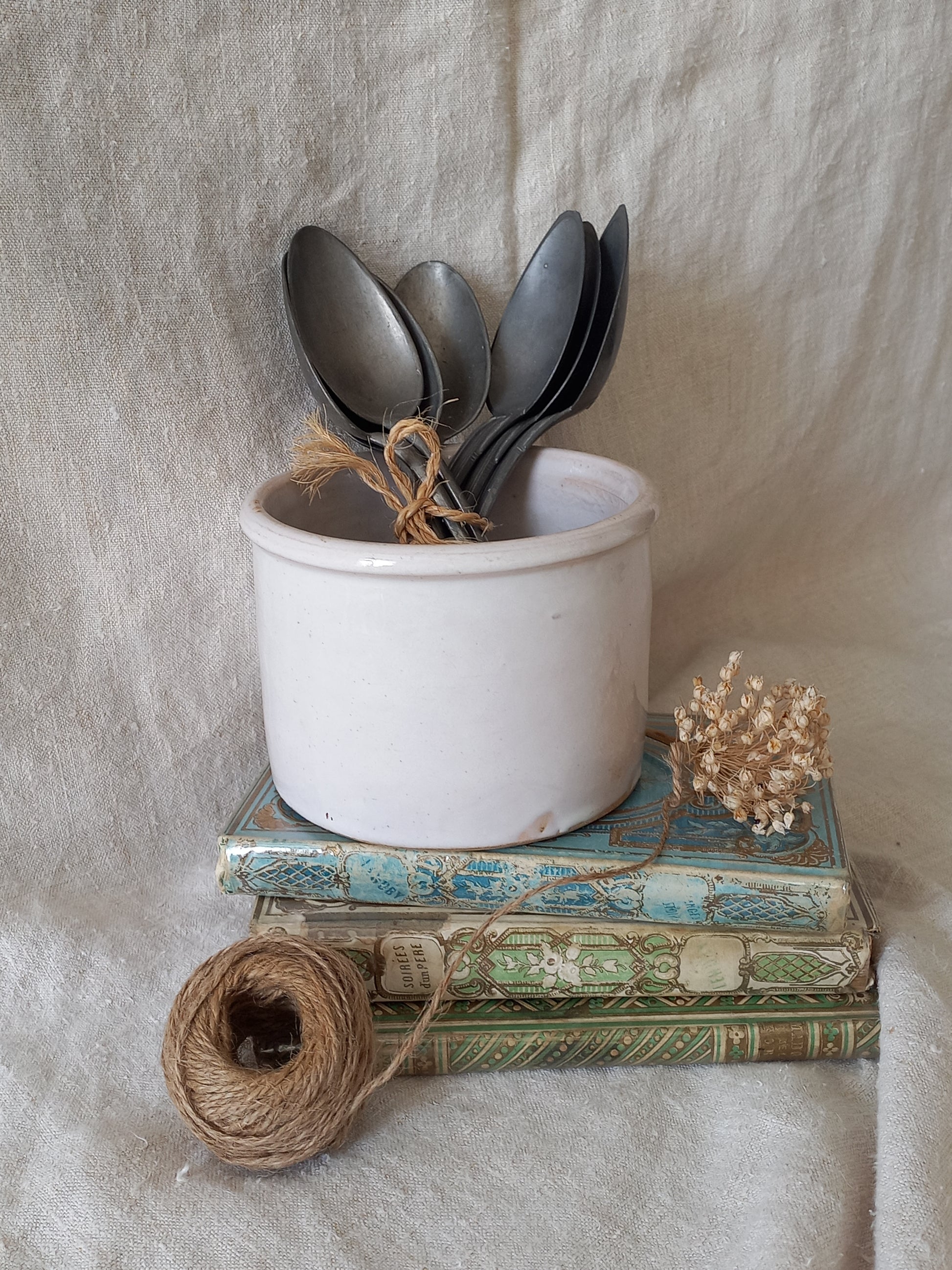 Antique jam pot on a textured surface containing spoons, dried flowers, and books on a textured surface