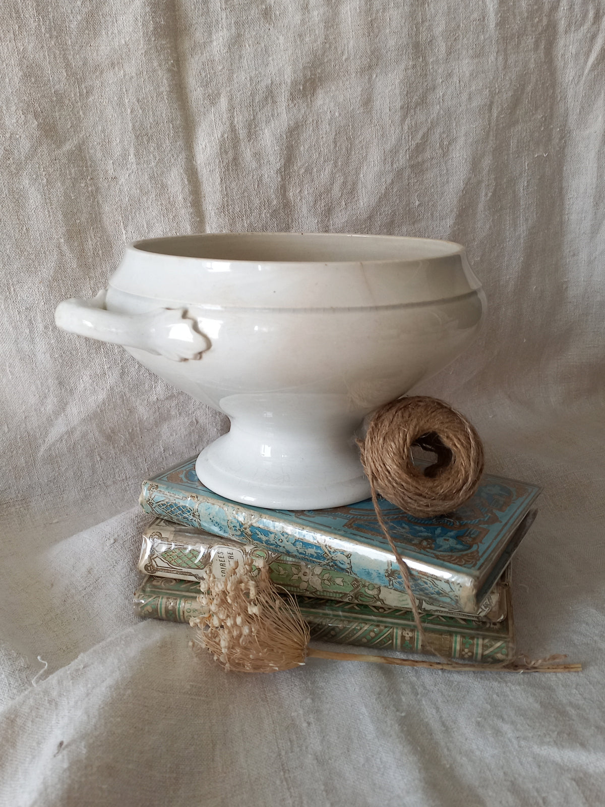 White ironstone bowl on a stack of books with twine and dried flowers against a textured fabric background