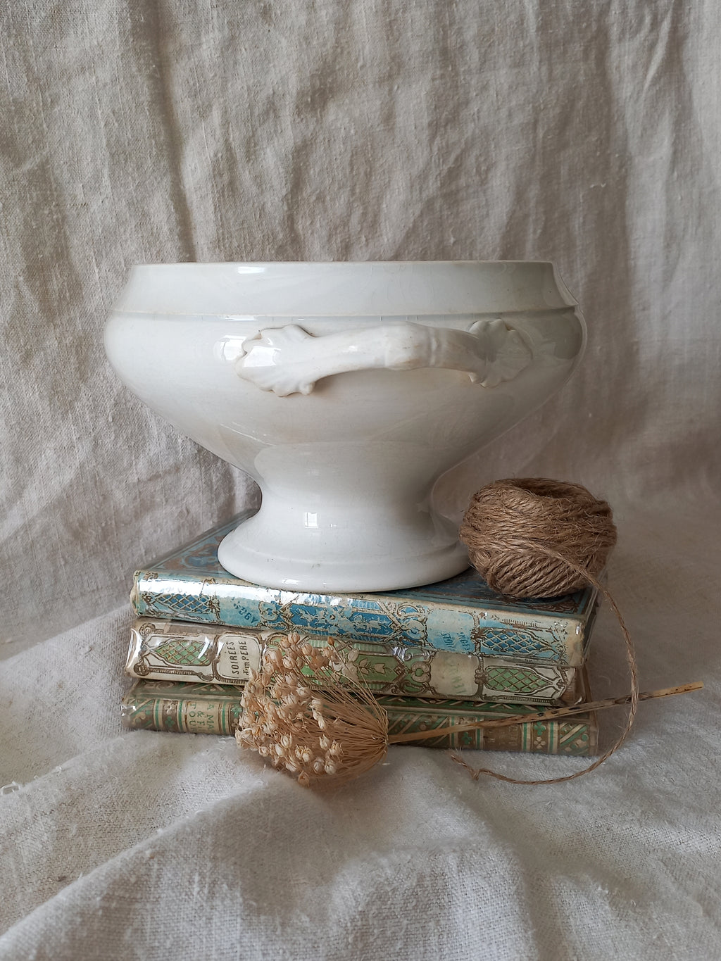 White ironstone bowl on a stack of books with twine and dried flowers against a textured fabric background