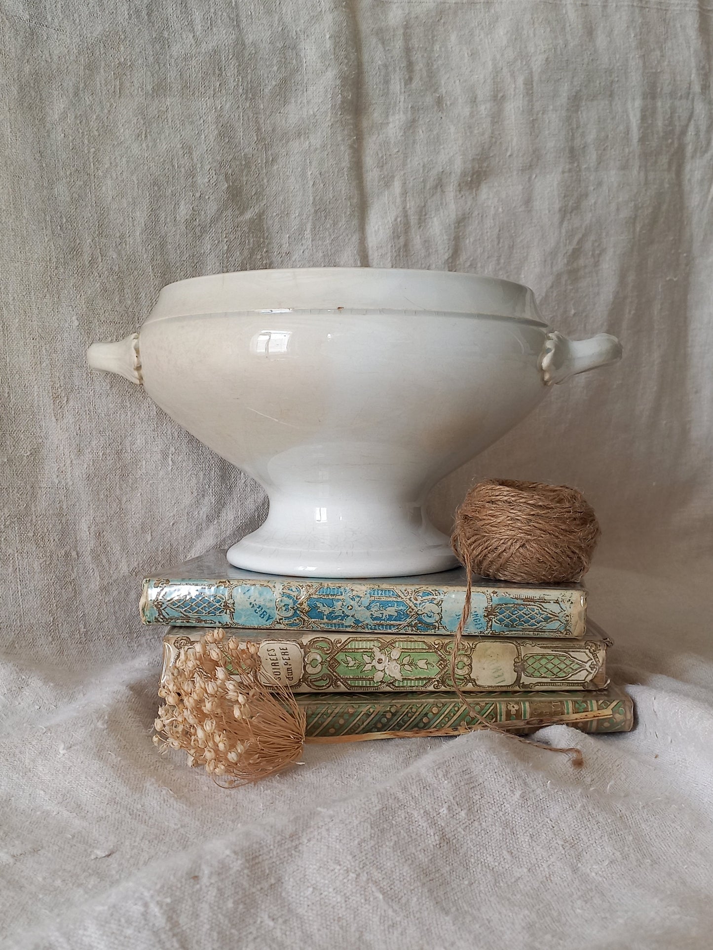White ironstone bowl on a stack of books with twine and dried flowers against a textured fabric background