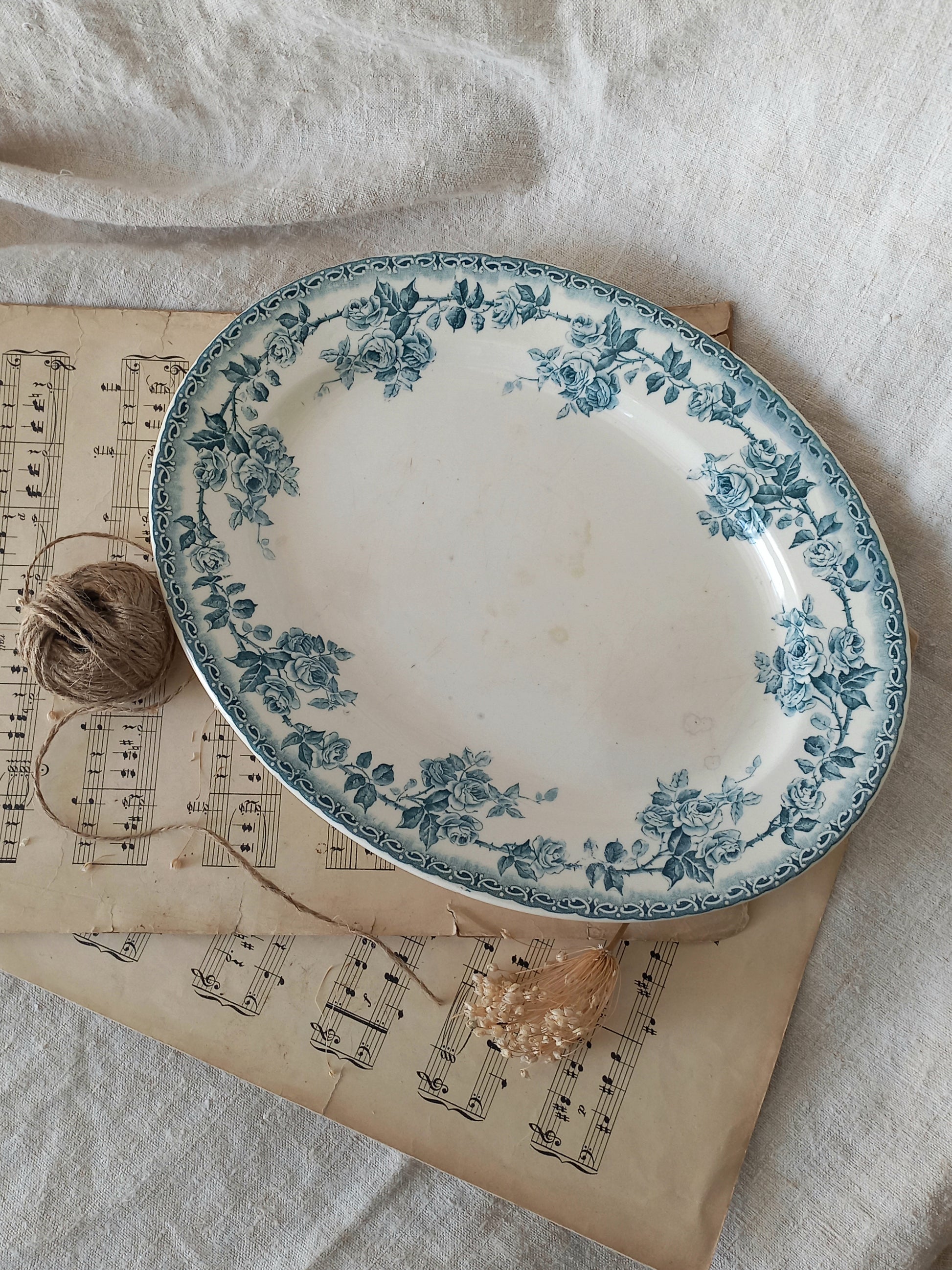 An oval ceramic plate with a blue floral border pattern, styled on top of vintage sheet music with a ball of twine and dried flowers on a white linen background.