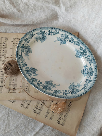 An oval ceramic plate with a blue floral border pattern, styled on top of vintage sheet music with a ball of twine and dried flowers on a white linen background.