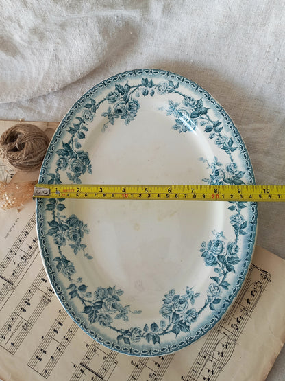 An oval white ceramic plate with a blue floral border pattern, shown with a yellow tape measure across the middle to indicate size. The plate is styled on top of vintage sheet music next to a spool of twine on a white fabric background.