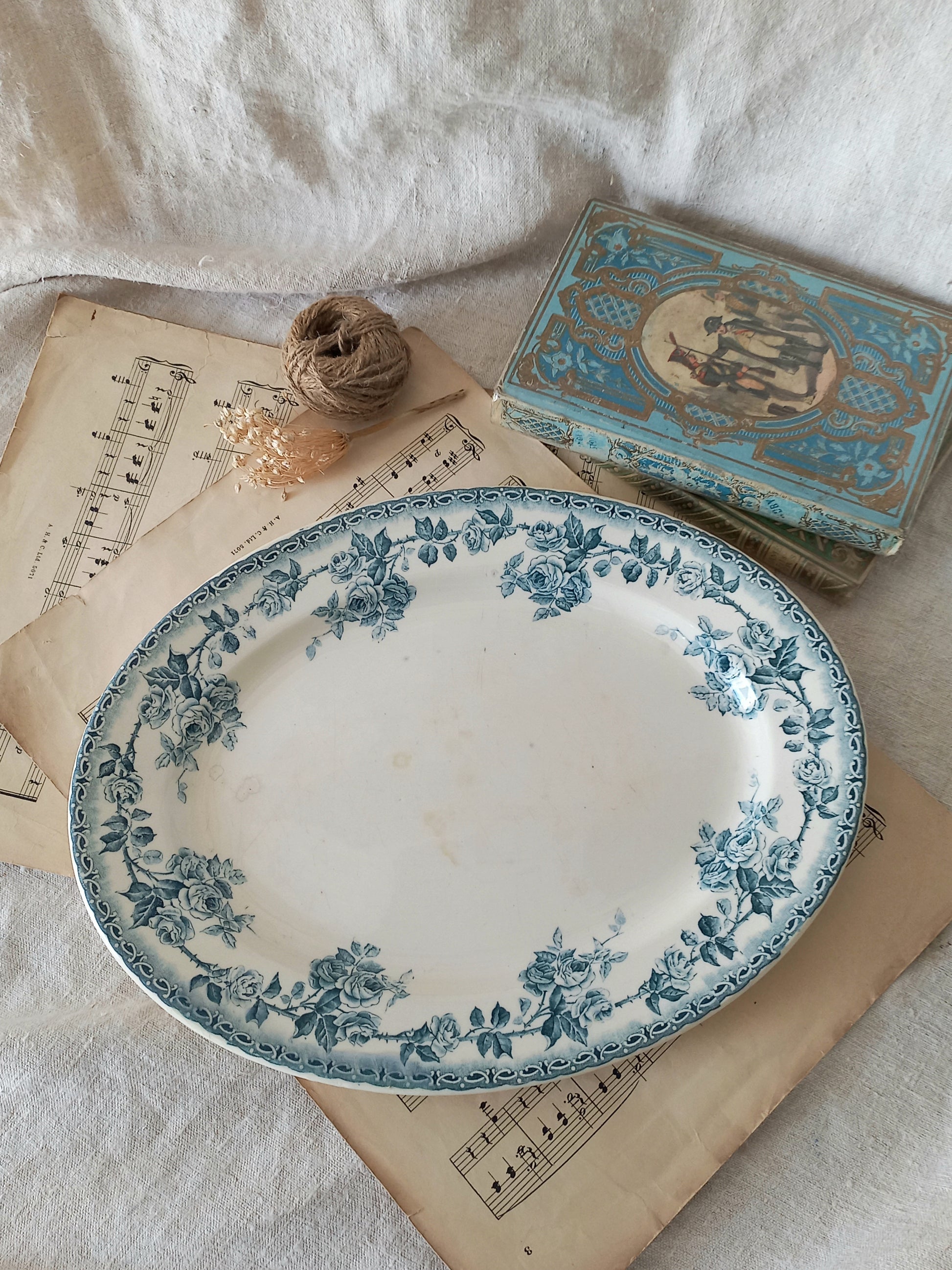 A vintage blue and white floral ceramic serving platter displayed on antique sheet music, next to a small decorative box and twine on a white fabric background.