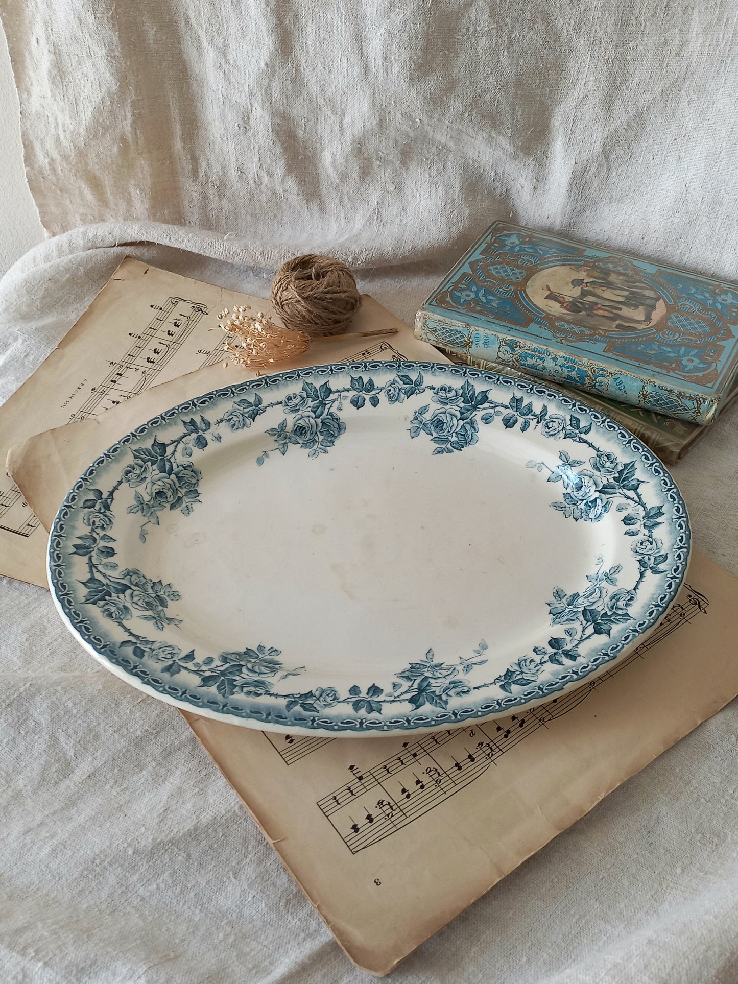 An oval ceramic serving platter with a blue floral border pattern on a white base, styled on vintage sheet music with a small book and twine against a draped white fabric background.