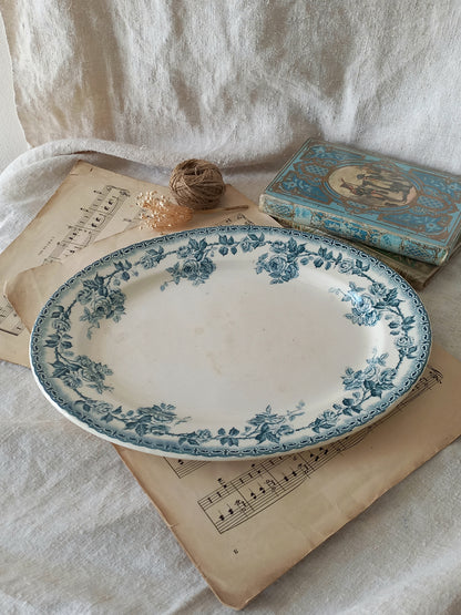 An oval ceramic serving platter with a blue floral border pattern on a white base, styled on vintage sheet music with a small book and twine against a draped white fabric background.