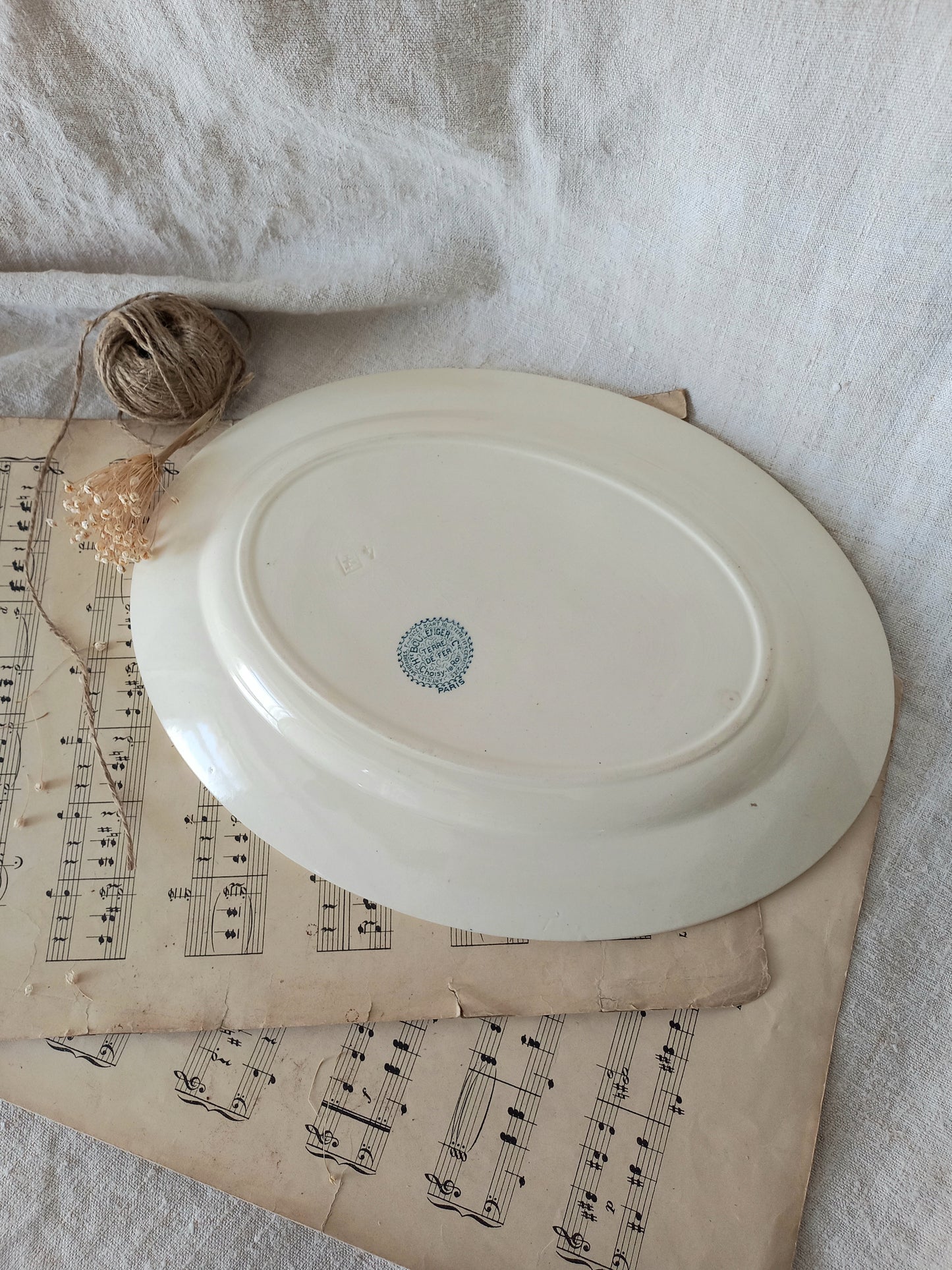An off-white ceramic oval plate with a small blue circular stamp in the center, styled on top of vintage sheet music with a small ball of twine and dried flower against a white fabric backdrop.