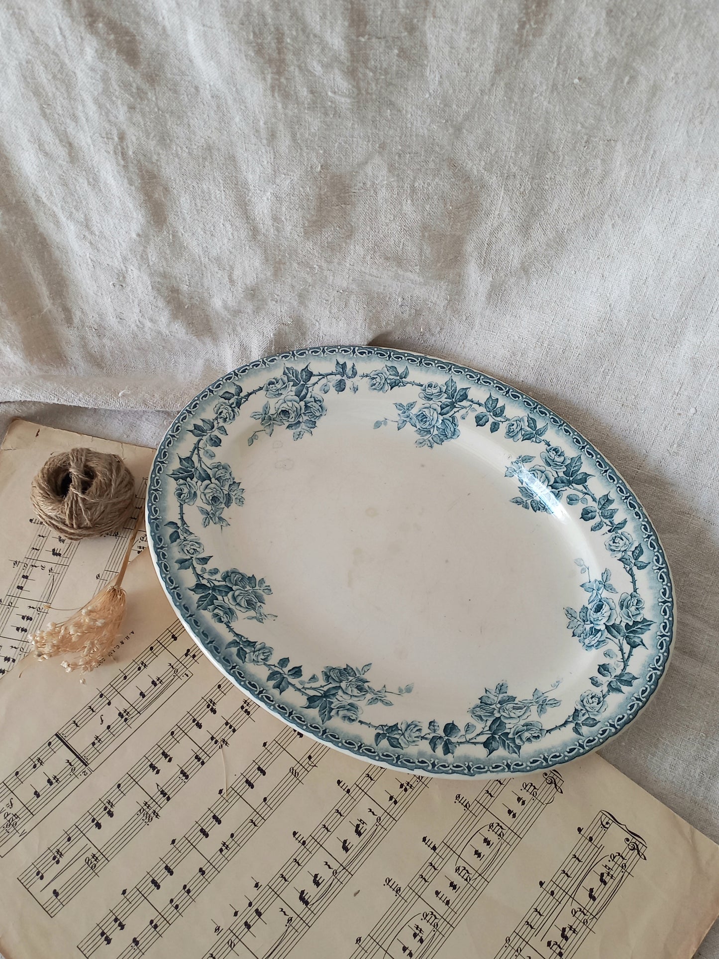 An oval ceramic plate with a blue floral toile pattern on a white background, styled on vintage sheet music next to a small spool of twine and a dried flower against a draped white fabric backdrop.
