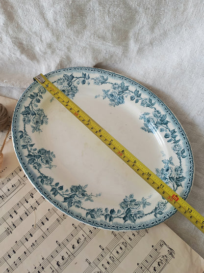 An oval ceramic serving platter with a blue floral pattern on a white background, shown with a yellow tape measure laid diagonally across it on a white fabric surface with sheet music underneath.