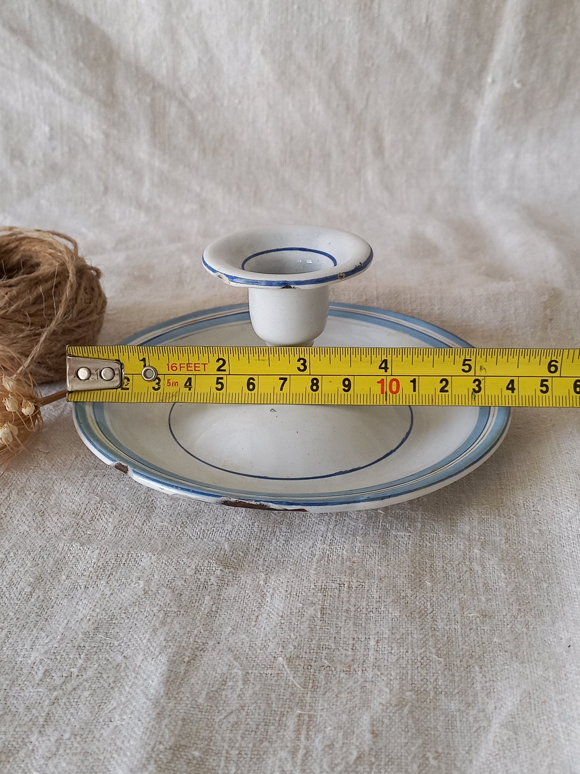 White enamel candle holder with blue rim on a textured beige surface with twine and dried flowers and a tape-measure for reference.