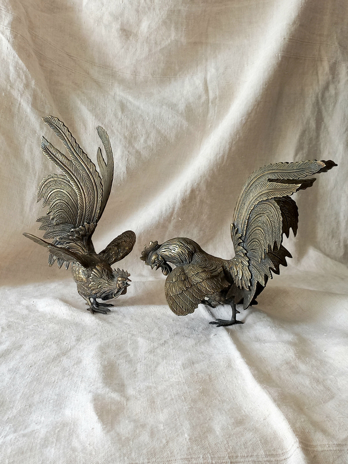 Two metallic rooster sculptures on a textured white fabric background