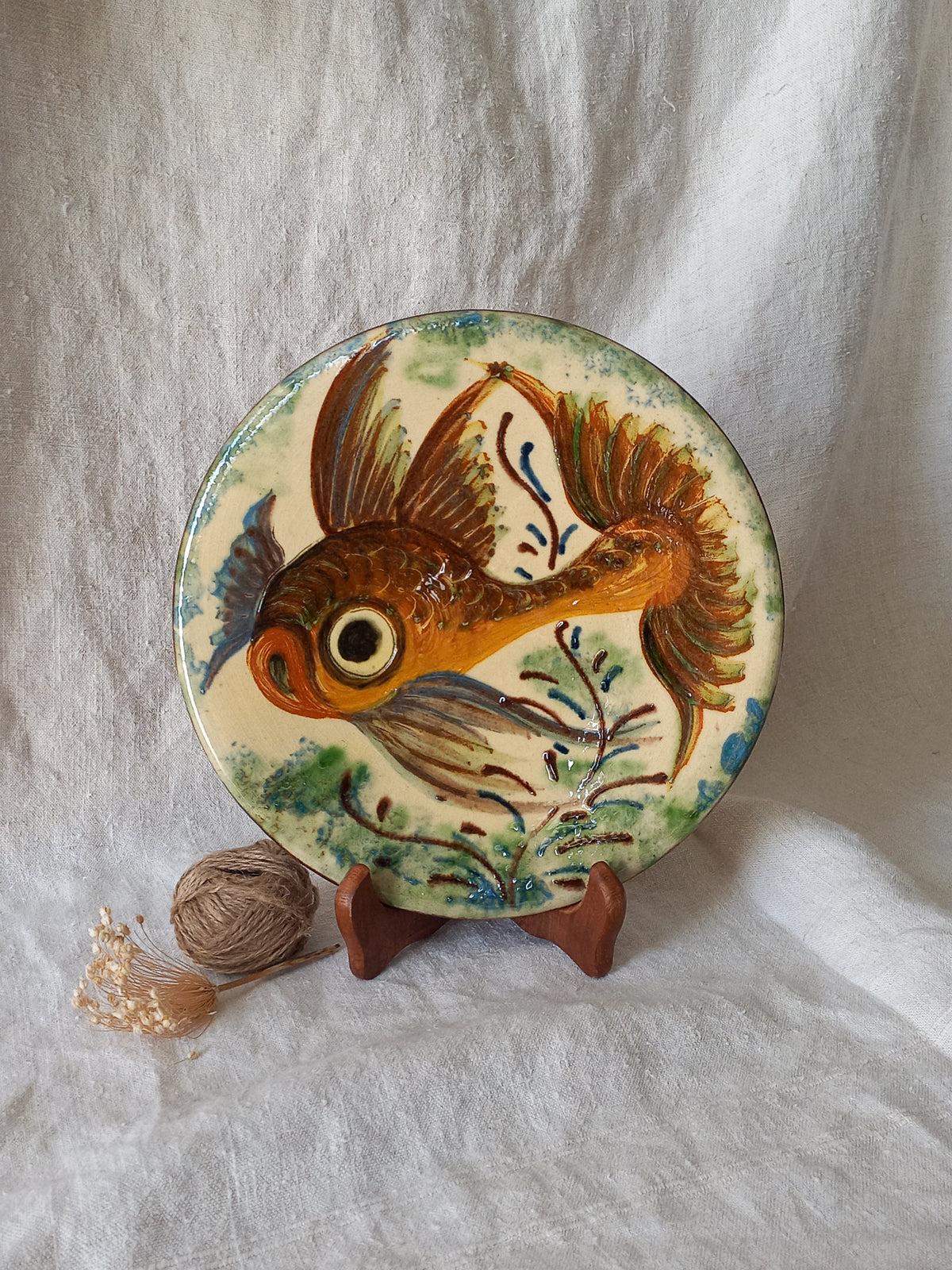 A round ceramic plate featuring a hand-painted goldfish design, displayed on a small wooden stand against a draped white fabric background.