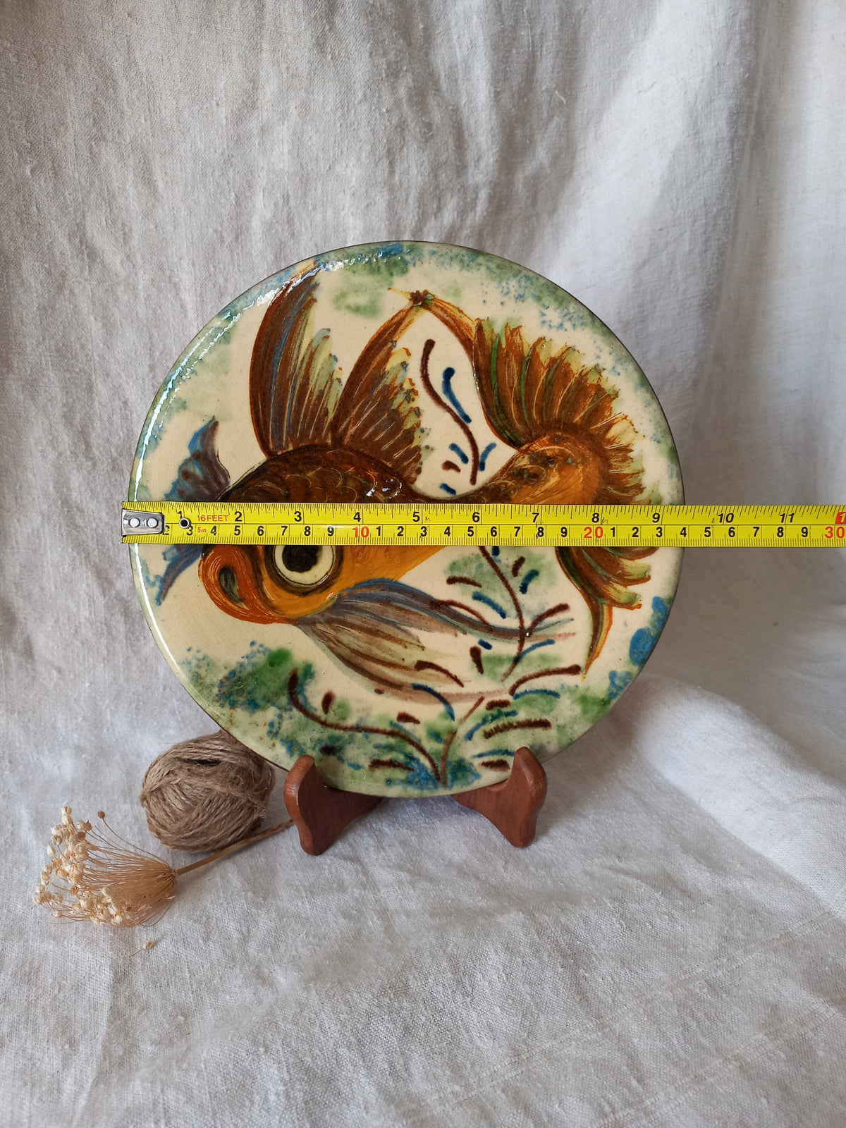 A round ceramic plate with a hand-painted goldfish design, displayed on a small wooden stand with a yellow tape measure across the middle to show its diameter, set against a draped white fabric background.