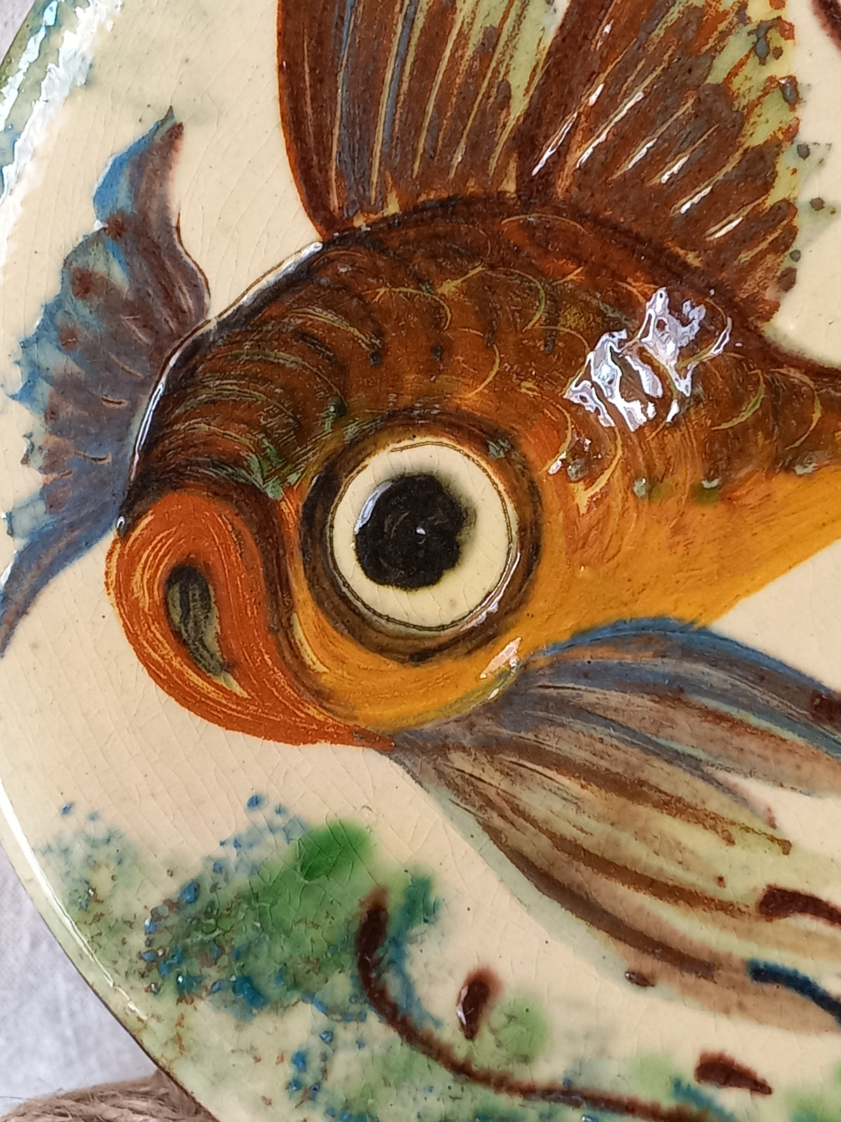Close-up of a hand-painted ceramic fish, likely a goldfish, featuring brown, orange, blue, and green glazes on a cream-colored plate.