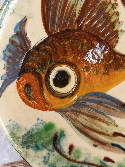 Close-up of a hand-painted ceramic fish, likely a goldfish, featuring brown, orange, blue, and green glazes on a cream-colored plate.