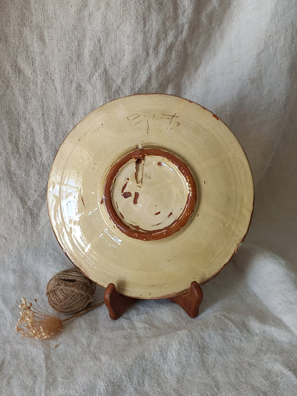 Rear of a round ceramic plate with a glossy cream glaze and a rustic reddish-brown rim, displayed on a small wooden stand against a draped white fabric background.