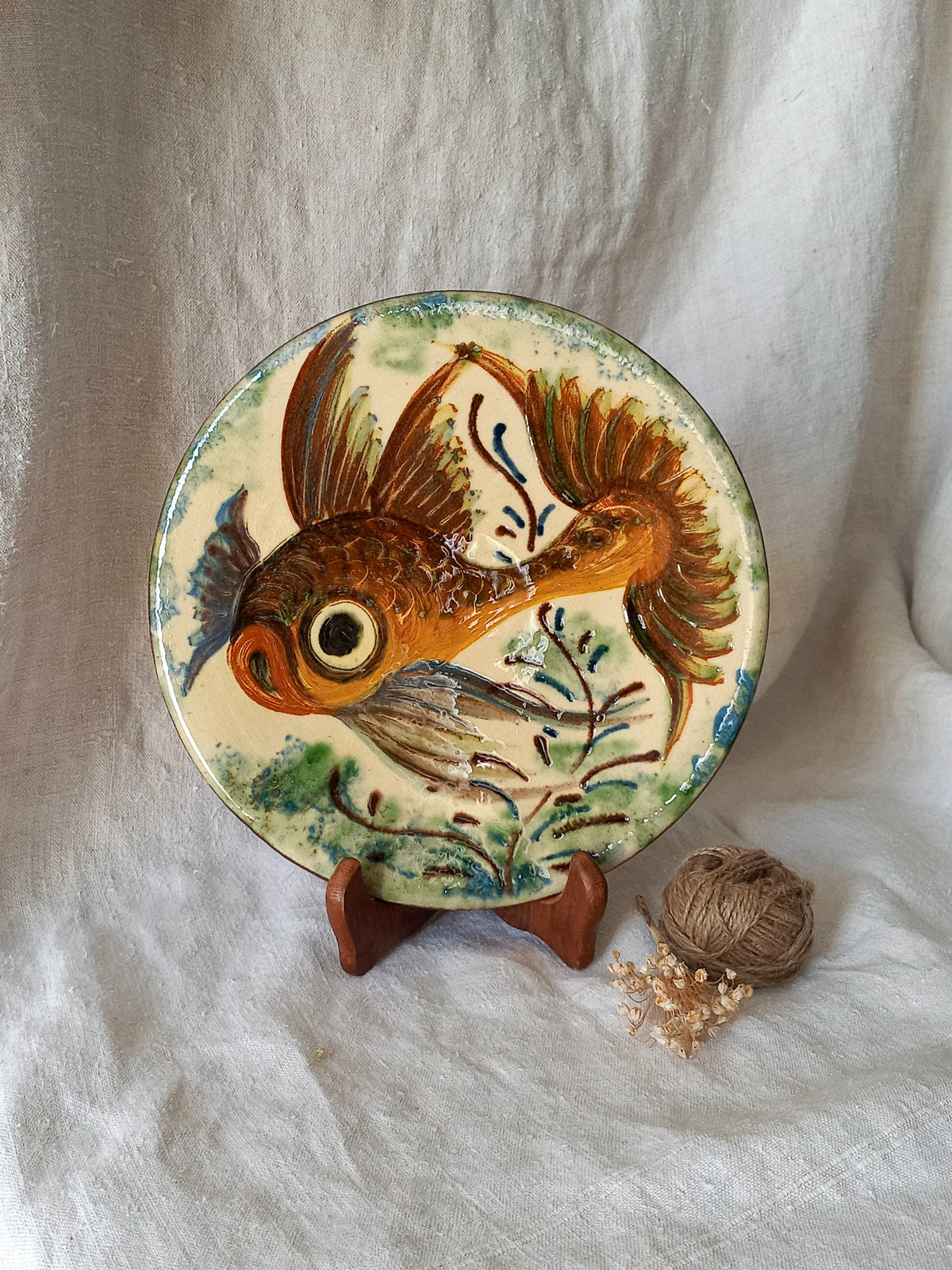 A round ceramic decorative plate featuring a hand-painted goldfish design, displayed on a small wooden stand against a draped white fabric backdrop.