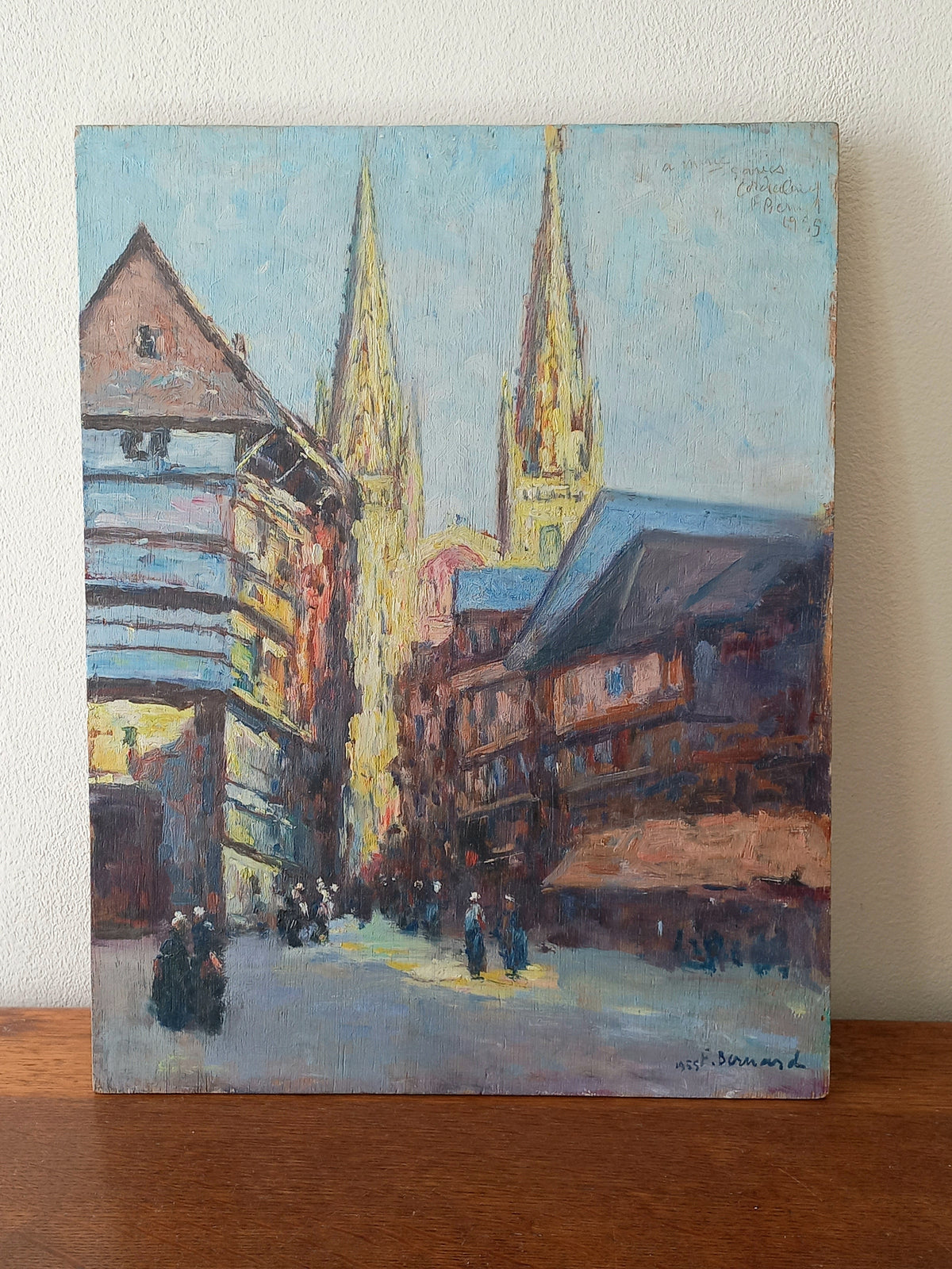An impressionist-style painting of a European street with two tall church spires, housed in buildings, displayed on a wooden surface against a light wall.