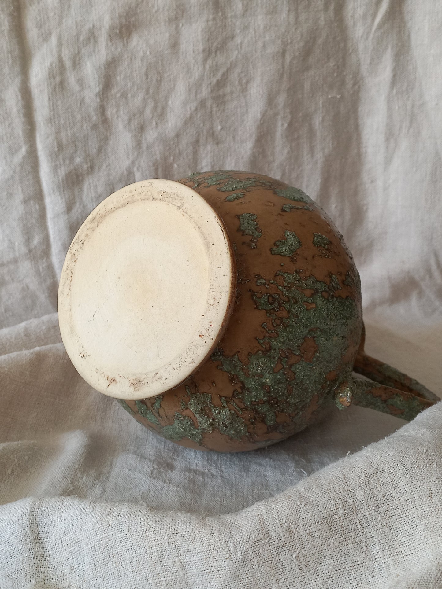 Underneath of a large vintage pottery jug with green and earth tones on a beige background